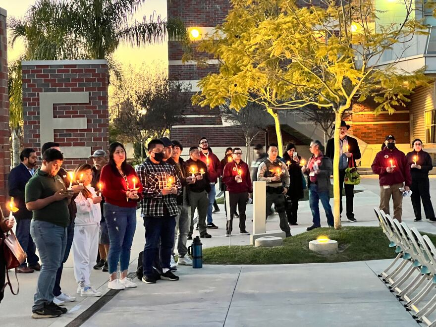 A group gathers around a tree in a campus quad holding candles. The trees are uplit as it is evening and a brick column with a large E on it