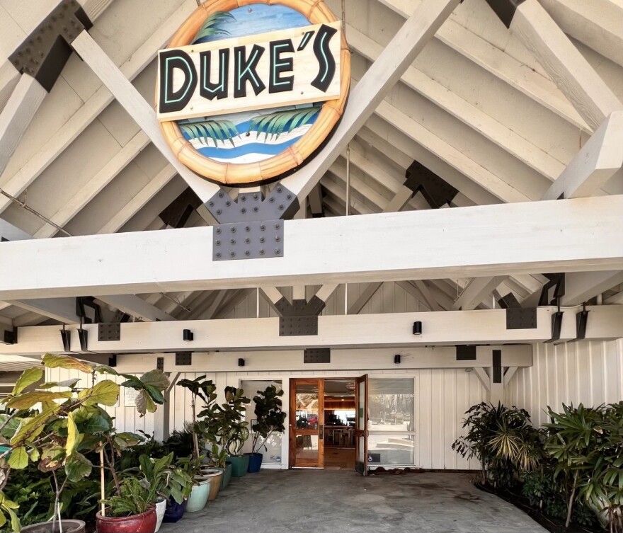 Outdoor entrance to Duke's restaurant with potted plants lining the walkway and a large wooden sign reading "DUKE'S" above the doorway.
