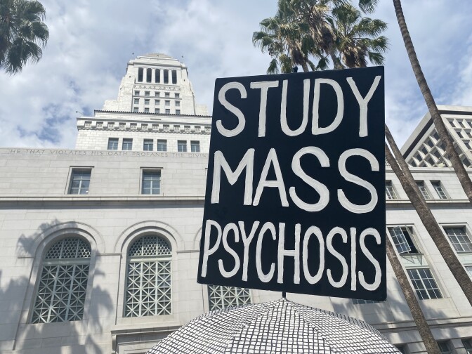 A sign from the anti-vaccination rally in downtown LA on Aug. 14, 2021. A sign with black background and white lettering that reads in all capital letters, "study mass psychosis" is seen in the foreground, with LA city hall looming in the background against a partly cloudy sky.