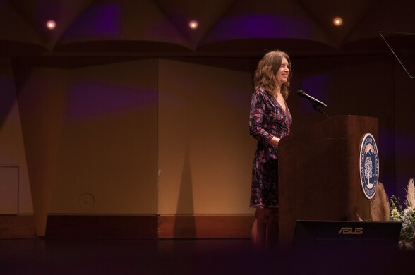 Person with light skin and long hair and wearing a dress speaks at podium.