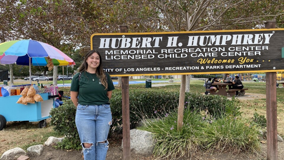 A Latina wearing a green shirt and jeans stands in front of a sign for the Hubert H. Humphrey Park. 