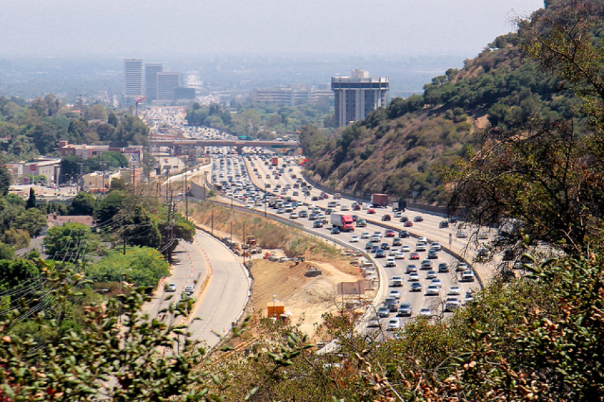 An aerial view of a curving freeway with five lanes going each direction and brush on either side.