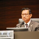 A man in a chair wearing a suit jacket, tie and glasses looks forward with a microphone in front of him. A sign in front has the official seal of the County of Orange and states "Andrew Do, Vice Chairman, District 1."