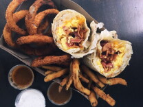 onion rings, breaded and fried zucchini sticks and a breakfast burrito filled with eggs, bacon and cheese sit in a brown cardboard box