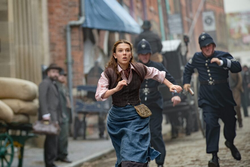 Millie Bobby Brown is in the center of the image mid run. She wears a faded blue skirt, a brown button up vest, and a light pink collared shirt underneath. The shirt sleeves are rolled up to her elbows and the skirt is gathered at the waist to shorten the hem length. Around her waist is also a small yellow pouch that peeks out from her vest. Brown's left arm is in front of her, slightly blocking her chest, while her right arm stretches behind her. Her arms are pumping alongside her as she sprints through a dirt street. Behind her, seemingly chasing after her, are two police officers. Their uniforms are from the 1800s (the time period of the film), with navy blue coats that hit mid-thigh with brass round buttons up the middle, and matching navy blue pants. The two officers also wear helmets, almost egg-shaped, with straps across their chins to help keep them in place. One of the officers on the left is partially blocked by Brown's frame, while the other officer on the right can be fully seen, his left hand holding a cylindrical tan item. In the background along the street are men in suits watching next to carts full of burlap sacks. A horse drawn carriage can be seen behind the two officers as well as signs for storefronts to the right, indicating a bustling city. 