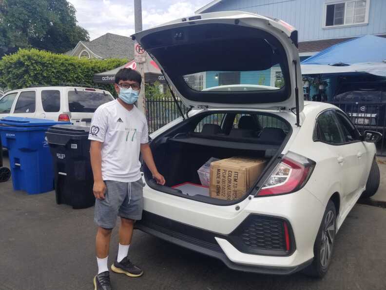 A man wearing a white shirt, grey pants and a face mask stands at the back of his white car, the hatch open, with a box inside the hatch.