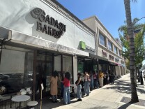 A wide shot of people lined up outside a bakery on a sunny day in Pasadena. The bakery sign reads on a white building "BAD ASH BAKES"