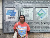 A teenager with her hair pulled back in a ponytail stands in front of a small building. She smiles at the camera. A sign on the wall behind her lists the agencies and politicians involved in transforming the space into a park. 