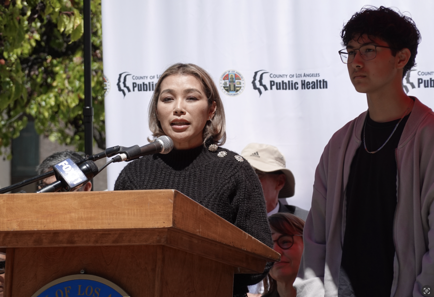 A woman speaks into a microphone on a podium as a teenage boy wearing glasses stands behind her.