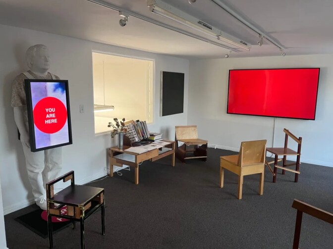 a room with wooden chairs and a red screen on the wall and a white statue of a man holding a sign that says "you are here" in white text on a red circle