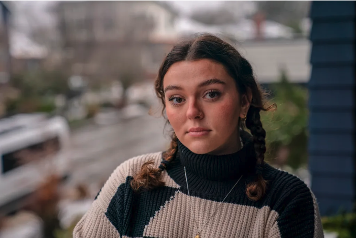 A woman who appears to be white and in her 20s is shown from the shoulders up. She looks pensively at the camera. Her hair is brown and worn in two braids. She wears a black and tan turtleneck sweater and silver necklace. She is outdoors and a blurry street can be seen in the background. It is day time.