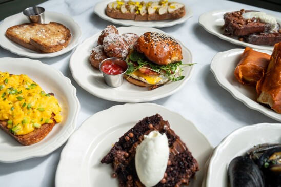 A spread of several prepared dishes displayed on a table with a white cloth , including scrambled eggs on toast, a breakfast sandwich with potatoes, chocolate, and oysters. 