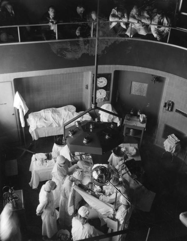 People stand on a balcony overlooking a medical procedure in progress below. Doctors stand working under a lighting device. They wear white scrubs and head coverings.