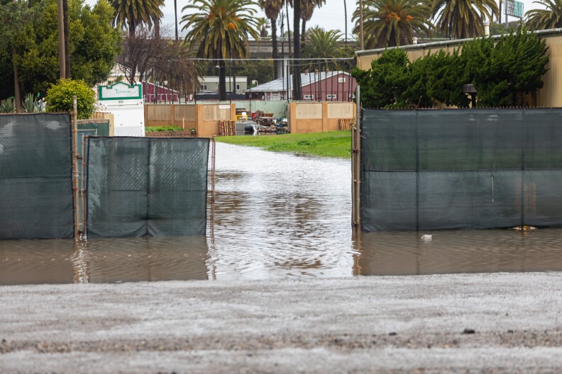 A an opening in a metal gate with black tarp reveals a flooded passageway next to a green lawn. In the background there are structures and palm trees.