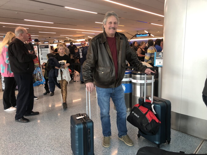 A man smiles and wears a brown jacket, maroon shirt and jeans with each hand on the handle of a suitcase. A crowd of people are behind him.