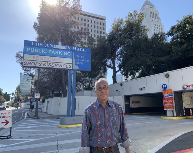 A Chinese American man in his early 70s, who's wearing glasses and a bright, printed shirt, stands in front of the entrance of a parking garage.
