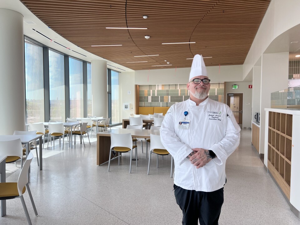 A white man wearing chef whites stands in a white and beige dining room. 