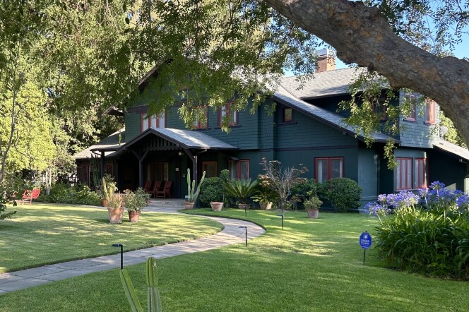 A dark green house with red window paneling is surrounded by bushes. There is a green front lawn and a cement path leads to the home's front door. A large tree sits out front. 
