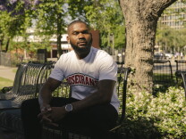 A man with a beard, white shirt that says "Rosecrans," and black pants sits on a bench among trees and jasmine at a college campus. 