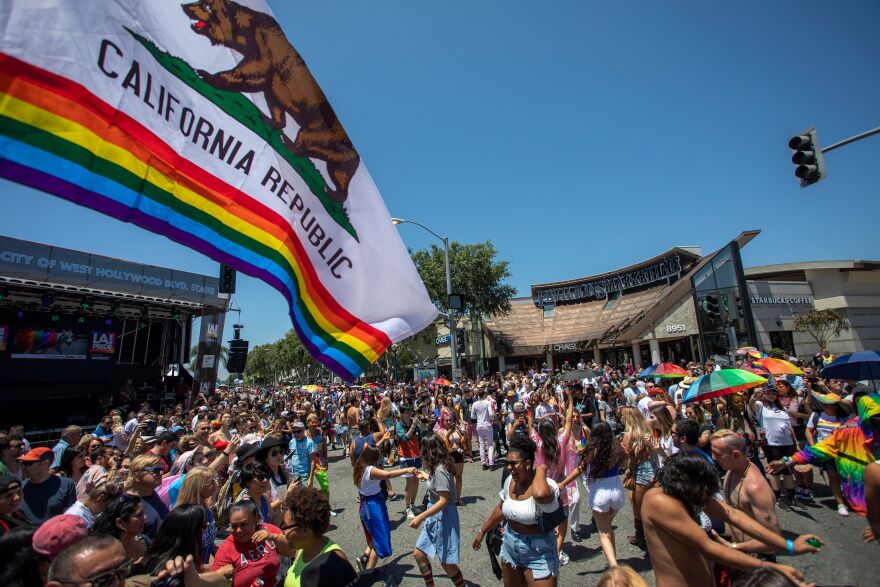An eclectic crowd of people gather on the concrete, some dancing, others cheering, and others with their arms thrown in the air. In the foreground waves a large California state flag--except its also partially a pride flag with the colors of the rainbow on the bottom half. A sunny cloudless sky hangs over the crowd's heads.