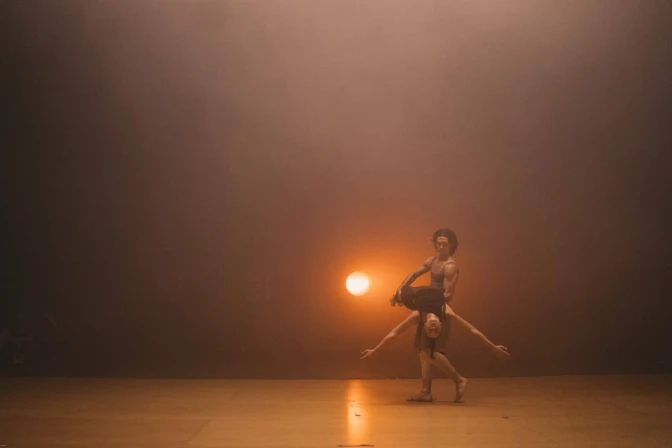A man holds a woman sideways with her arms out on a dance stage with a single spotlight behind them. 