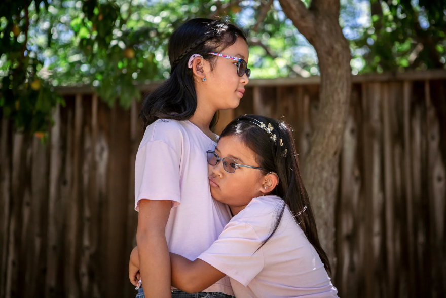 Two girls wearing hearing aids embrace in front of a wood fence.
