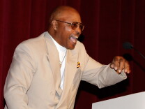 Actor Roger Mosley, a 50- or 60-something Black man in this image, is shown with an animated smile while speaking behind a podium. He wears a sports jacket, white mock turtleneck, gold chain and aviator sunglasses. 
