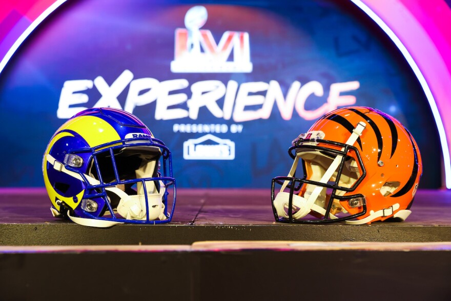 A blue-and-yellow Los Angeles Rams helmet faces opposite an orange-and-black Cincinnati Bengals helmet with the Super Bowl Experience logo as a backdrop.