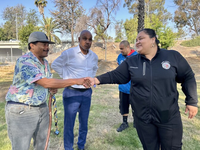 Two people shake hands. One is a female presenting person dressed in black. The other is a male presenting person wearing a cap.