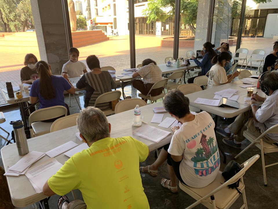A group of more than a dozen Japanese American volunteers of different ages sit at desks in a sunny room writing letters in support of reparations for Black Americans.