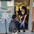 A woman in a black shirt and medium-tone skin pinky shakes a boy wearing a large gray backpack. A door shows different cartoon images of greetings like a hug, wave and fist pump. 