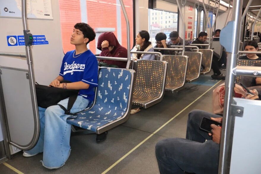 A high school aged student sits inside a metro train wearing a Dodgers jersey and blue jeans. There is a black backpack on his lap. Other people are seen on the train behind him.
