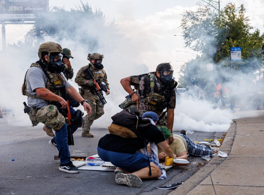 Federal officers wearing gas masks and holding weapons detain and approach a person on the floor as smoke fills the area of a street during the daytime.
