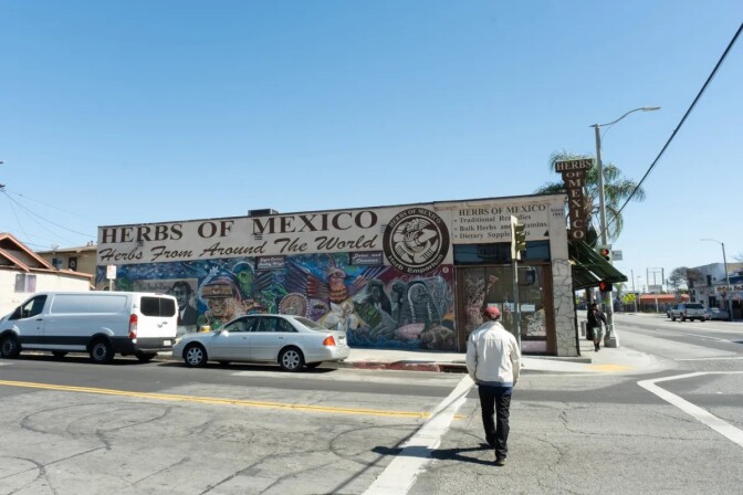 A man wearing a beige jacket, dark pants and a red baseball cap crossing a street. He is walking towards a building with a mural of people painted on the side with the words "herbs of Mexico. Herbs from around the world."