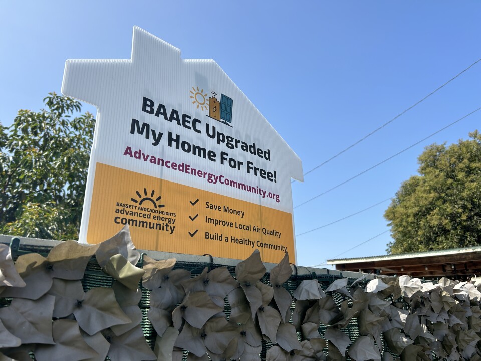 A sign that reads "BAAEC Upgraded My Home For Free! AdvancedEnergyCommunity.org" among other things on the top of a fence. The sign is shaped like a single-family house. 