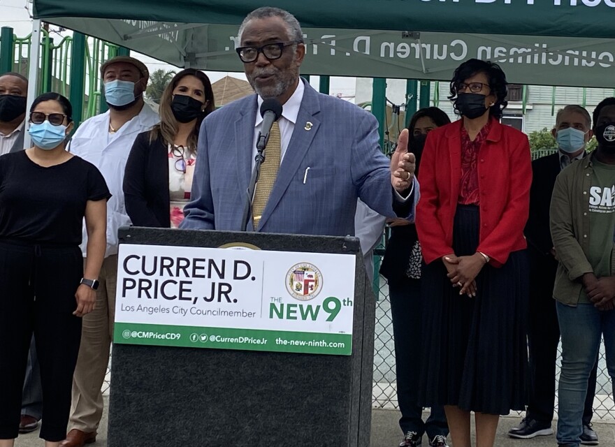 L.A. City Councilmember Curren Price, wearing a blue suit, stands at a podium with a sign attached to it that includes his name and a City Council, during an outdoor press conference. Several people wearing masks and standing under pop-up tent emblazoned with his name stand behind him.