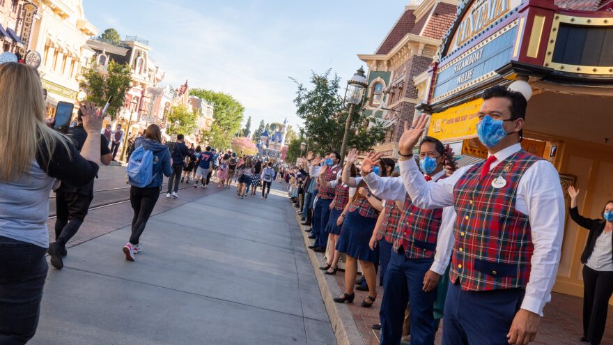 A line of Disney staff in sky blue face masks, plaid vests, and blue skirts or pants wave at people walking down Main Street U.S.A. at Disneyland.
