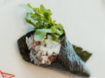 A hand roll wrapped in nori, filled with rice, dry-aged beef, melted cheese with visible char marks, and topped with a bundle of fresh arugula, photographed close-up against a white background.
