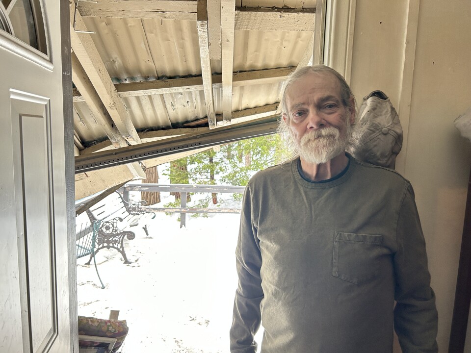 A man with light-tone skin and a short white beard stands in an open doorway. Behind him is a sagging eave and in the background a snow-covered deck.