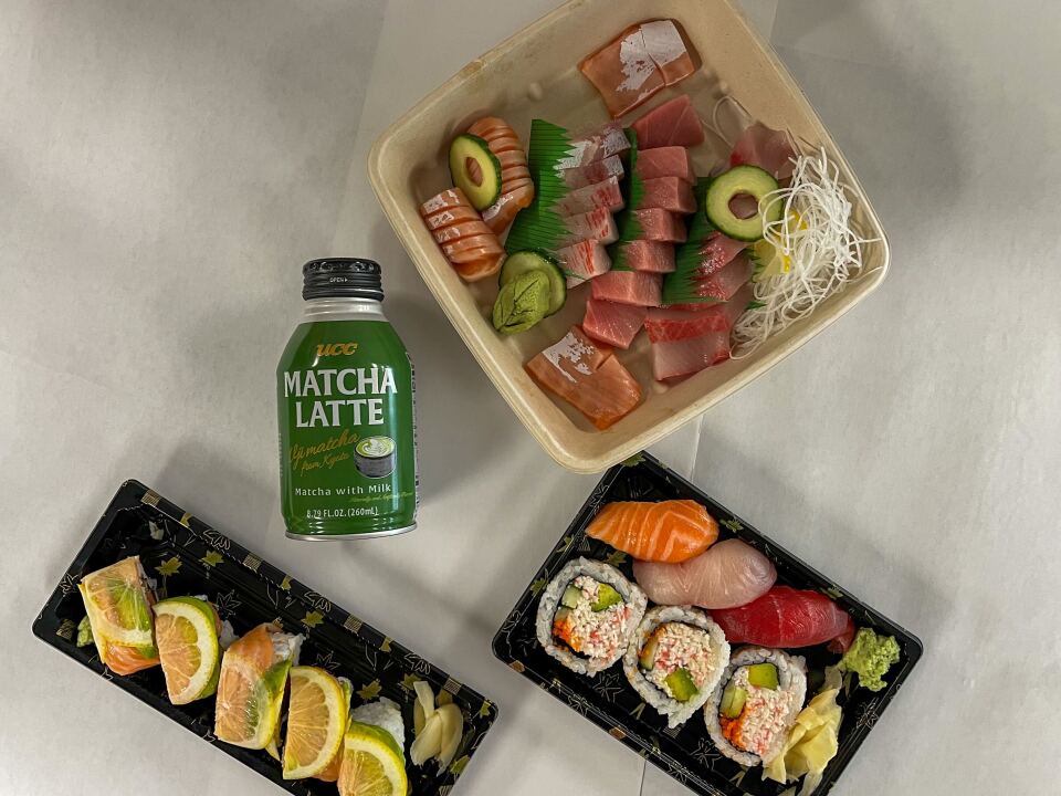 Three containers of sushi on a white background. On the bottom left is a roll containing thinly sliced lemon topped with pink salmon and avocado. The container next to it contains three large pieces of cut California roll sushi and pink salmon,  white escolar, and red tuna sushi. Above  is a large square container featuring sliced fish arranged decoratively. Between the containers is a short aluminum bottle with a green label and black top with the words 'Matcha Latte' printed on it.