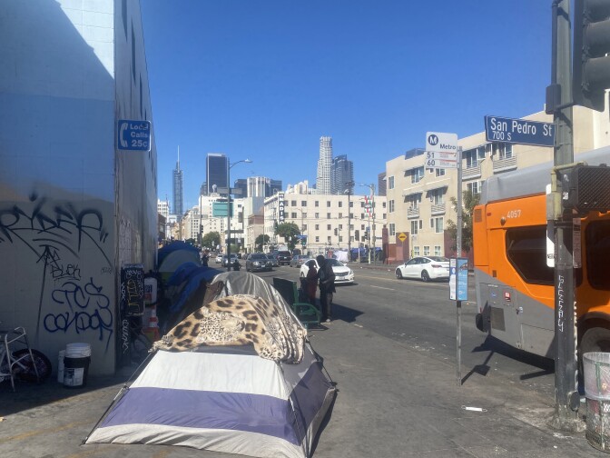 Tents line the sidewalk along a street on a sunny day as an orange and gray bus drives by. In the distance is the L.A. skyline.