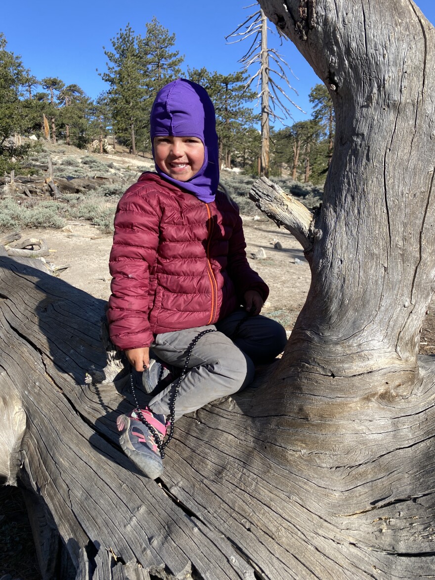A girl in a maroon-colored puffy jacket and purple balaclava sits on a tree trunk, smiling for the camera. 