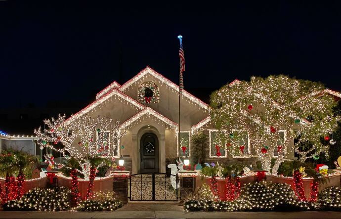 A home is decorated with white and red lights for the holidays. Two trees in front of the homes are also decorated with lights as are the bushes on the sidewalk. 