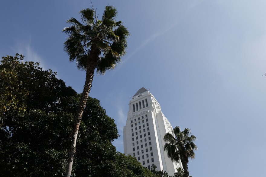 A view of Los Angeles City Hall from below, with a tall palm tree in the forefront and the light blue sky in the background.