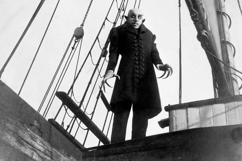 A black and white still of a vampire in a long black coat with claws and pointed ears, standing on a ship in the film "Nosferatu."