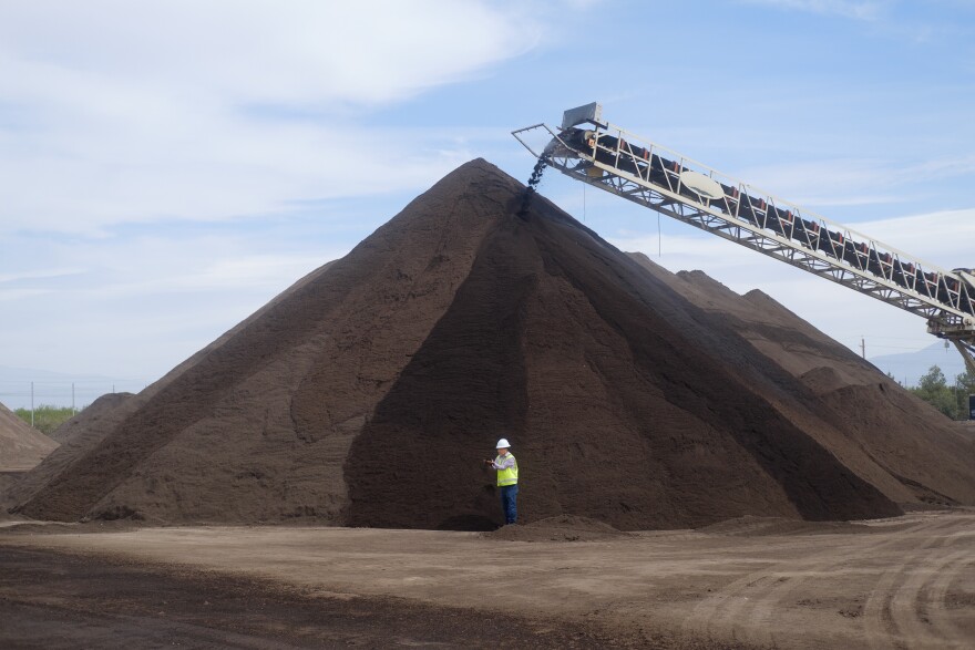A huge pile of dark brown compost in a dirt lot dwarfs a man wearing a white hard hat and neon safety vest.
