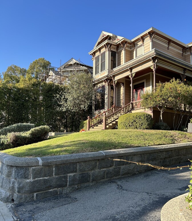 Looking up at a Victorian home in LA painted an ecru color with light green trim. It sits atop a big lawn surrounded by an old stone wall 