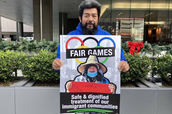 A man with a medium skin tone and black and gray hair wears a blue sweatshirt and holds a poster that reads "FAIR GAMES: Safe and dignified treatment of our immigrant communities."