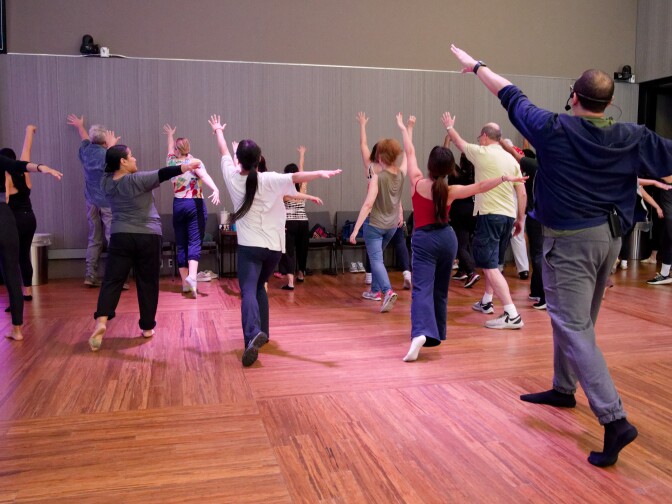 A wide shot of a group of people from behind, all standing on a wooden studio floor with their backs to the camera. They are following an instructor, who stands at the front right with a headset microphone, as they all reach one arm up and out in a synchronized movement toward the front of the room.