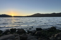 A large blue lake at sunset, with orange and yellow light peaking over a mountain range in the distance. 